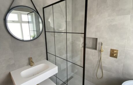 Contemporary bathroom designed by Hummingbird Bathrooms and Kitchens, featuring a round mirror above a white basin, glass shower partition with black frame, luxurious gold shower fittings, and grey tiled walls. Window reflection visible in the mirror. Specialising in bespoke bathrooms, kitchens, construction services and design in the UK. Contemporary bathroom designed by Hummingbird Bathrooms and Kitchens, featuring a round mirror above a white basin, glass shower partition with black frame, luxurious gold shower fittings, and grey tiled walls. Window reflection visible in the mirror. Specialising in bespoke bathrooms, kitchens, construction services and design in the UK.