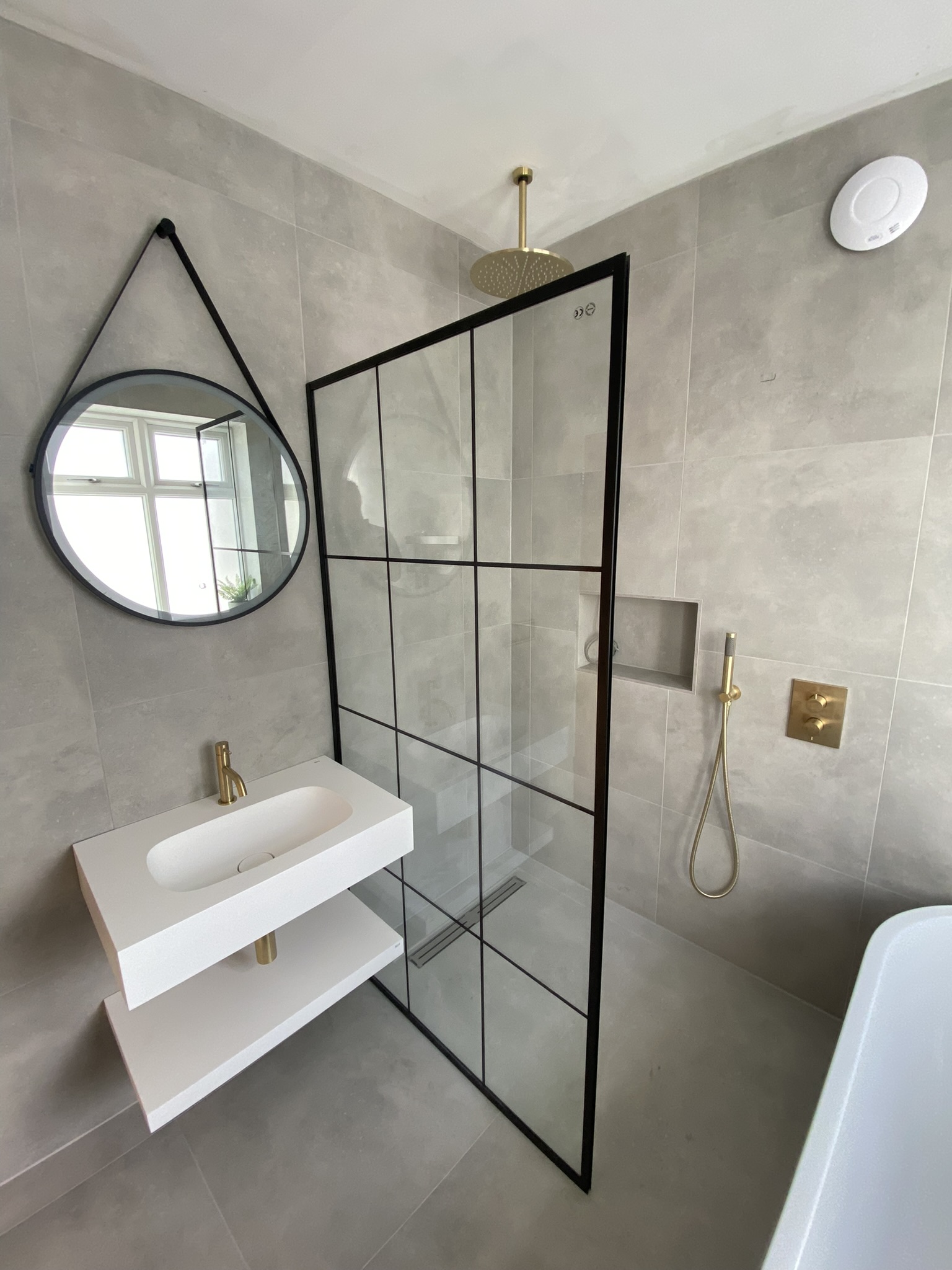 Contemporary bathroom designed by Hummingbird Bathrooms and Kitchens, featuring a round mirror above a white basin, glass shower partition with black frame, luxurious gold shower fittings, and grey tiled walls. Window reflection visible in the mirror. Specialising in bespoke bathrooms, kitchens, construction services and design in the UK. Contemporary bathroom designed by Hummingbird Bathrooms and Kitchens, featuring a round mirror above a white basin, glass shower partition with black frame, luxurious gold shower fittings, and grey tiled walls. Window reflection visible in the mirror. Specialising in bespoke bathrooms, kitchens, construction services and design in the UK.