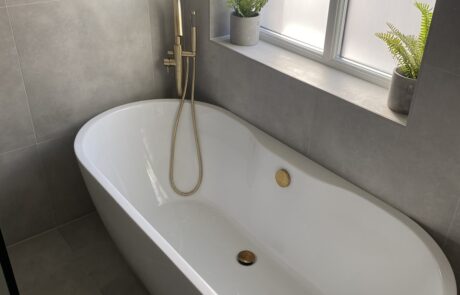 Modern bathroom design by Hummingbird Bathrooms and Kitchens featuring a white freestanding bath, elegant gold fittings, grey tiled walls and floor, and two small potted ferns on the windowsill; expert bathroom renovation and construction services in the UK. Modern bathroom design by Hummingbird Bathrooms and Kitchens featuring a white freestanding bath, elegant gold fittings, grey tiled walls and floor, and two small potted ferns on the windowsill; expert bathroom renovation and construction services in the UK.