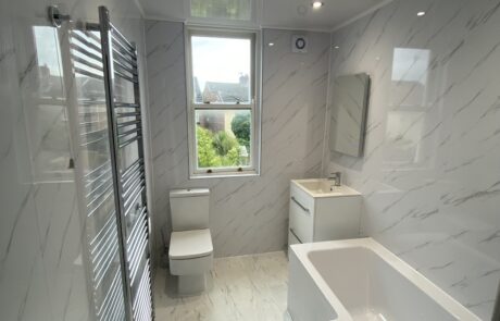 Contemporary bathroom design by Hummingbird Bathrooms and Kitchens featuring white marble walls and flooring, bath, toilet, basin with mirror, heated towel rail, and large window overlooking garden greenery. Bright space illuminated with recessed ceiling lights—premium bathroom renovation experts in the UK. Contemporary bathroom design by Hummingbird Bathrooms and Kitchens featuring white marble walls and flooring, bath, toilet, basin with mirror, heated towel rail, and large window overlooking garden greenery. Bright space illuminated with recessed ceiling lights—premium bathroom renovation experts in the UK.