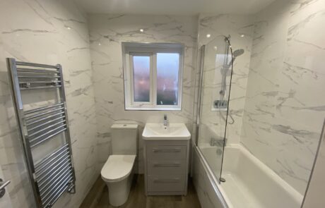 Hummingbird Bathrooms and Kitchens modern bathroom design featuring marble-style walls, a bath with integrated shower, contemporary washbasin unit, toilet, heated towel rail, and frosted window. Wood-effect flooring and ceiling spotlights complete the luxury bathroom refurbishment by our specialist UK team. Hummingbird Bathrooms and Kitchens modern bathroom design featuring marble-style walls, a bath with integrated shower, contemporary washbasin unit, toilet, heated towel rail, and frosted window. Wood-effect flooring and ceiling spotlights complete the luxury bathroom refurbishment by our specialist UK team.