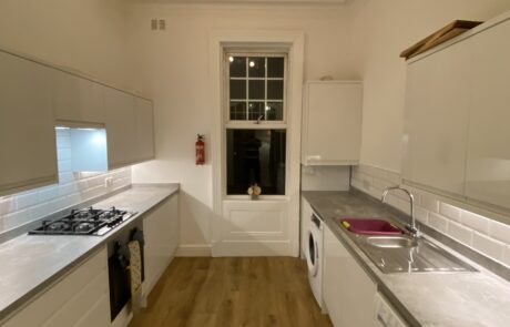 Contemporary kitchen by Hummingbird Bathrooms and Kitchens featuring wooden flooring, white cabinetry, integrated washing machines, modern gas hob, centred window with a mounted fire extinguisher, white tiled splashback, and triple ceiling lighting. Expert kitchen design and construction services in the UK. Contemporary kitchen by Hummingbird Bathrooms and Kitchens featuring wooden flooring, white cabinetry, integrated washing machines, modern gas hob, centred window with a mounted fire extinguisher, white tiled splashback, and triple ceiling lighting. Expert kitchen design and construction services in the UK.