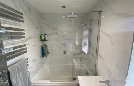 Modern bathroom by Hummingbird Bathrooms and Kitchens, featuring elegant marble-effect tiles, glass shower screen over a bath, chrome towel radiator on the left, white basin with silver tap on the right, and stylish blue and grey towels; expert bathroom design and installation in the UK. Modern bathroom by Hummingbird Bathrooms and Kitchens, featuring elegant marble-effect tiles, glass shower screen over a bath, chrome towel radiator on the left, white basin with silver tap on the right, and stylish blue and grey towels; expert bathroom design and installation in the UK.