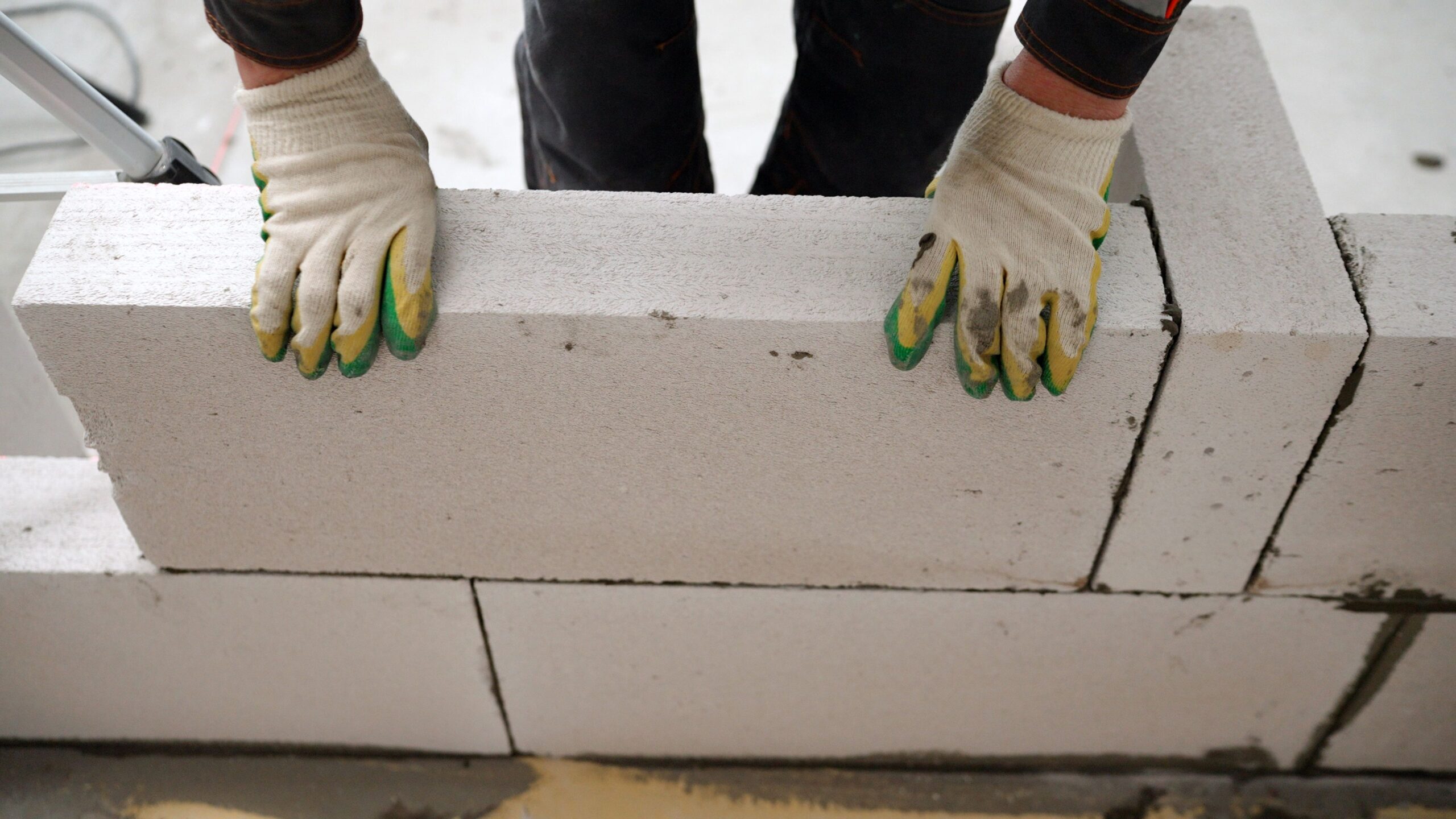 Builder wearing gloves carefully laying white concrete blocks with visible mortar lines, constructing a wall as part of Hummingbird Bathrooms and Kitchens’ expert bathroom, kitchen, and home design and construction services in the UK. Builder wearing gloves carefully laying white concrete blocks with visible mortar lines, constructing a wall as part of Hummingbird Bathrooms and Kitchens' expert bathroom, kitchen, and home design and construction services in the UK.