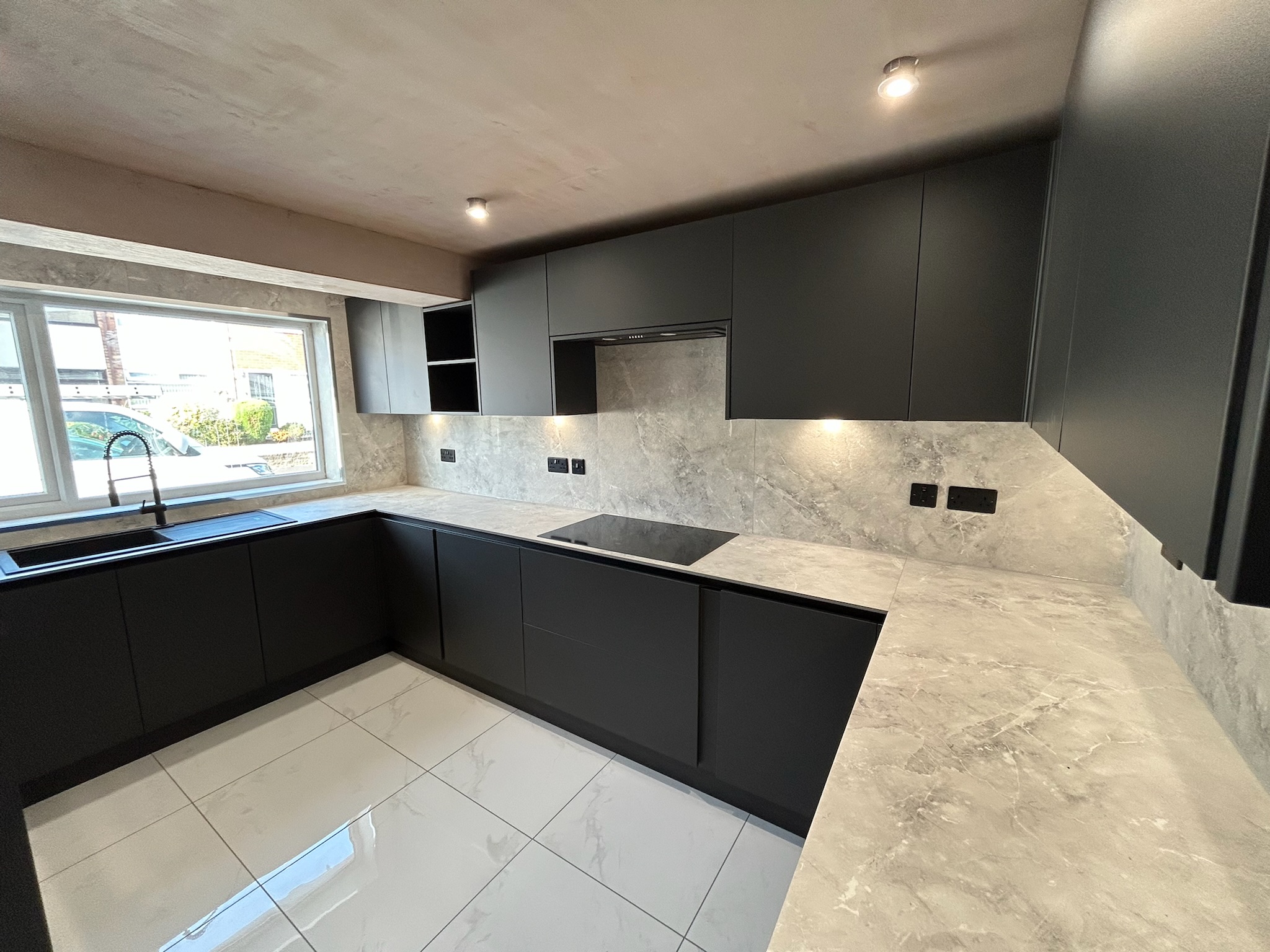 Contemporary kitchen designed by Hummingbird Bathrooms and Kitchens featuring matt black cupboards, light marble worktops, built-in hob, tiled flooring, under-cupboard lighting, and a large window above the sink providing natural daylight—expert kitchen design and installation services in the UK. Contemporary kitchen designed by Hummingbird Bathrooms and Kitchens featuring matt black cupboards, light marble worktops, built-in hob, tiled flooring, under-cupboard lighting, and a large window above the sink providing natural daylight—expert kitchen design and installation services in the UK.