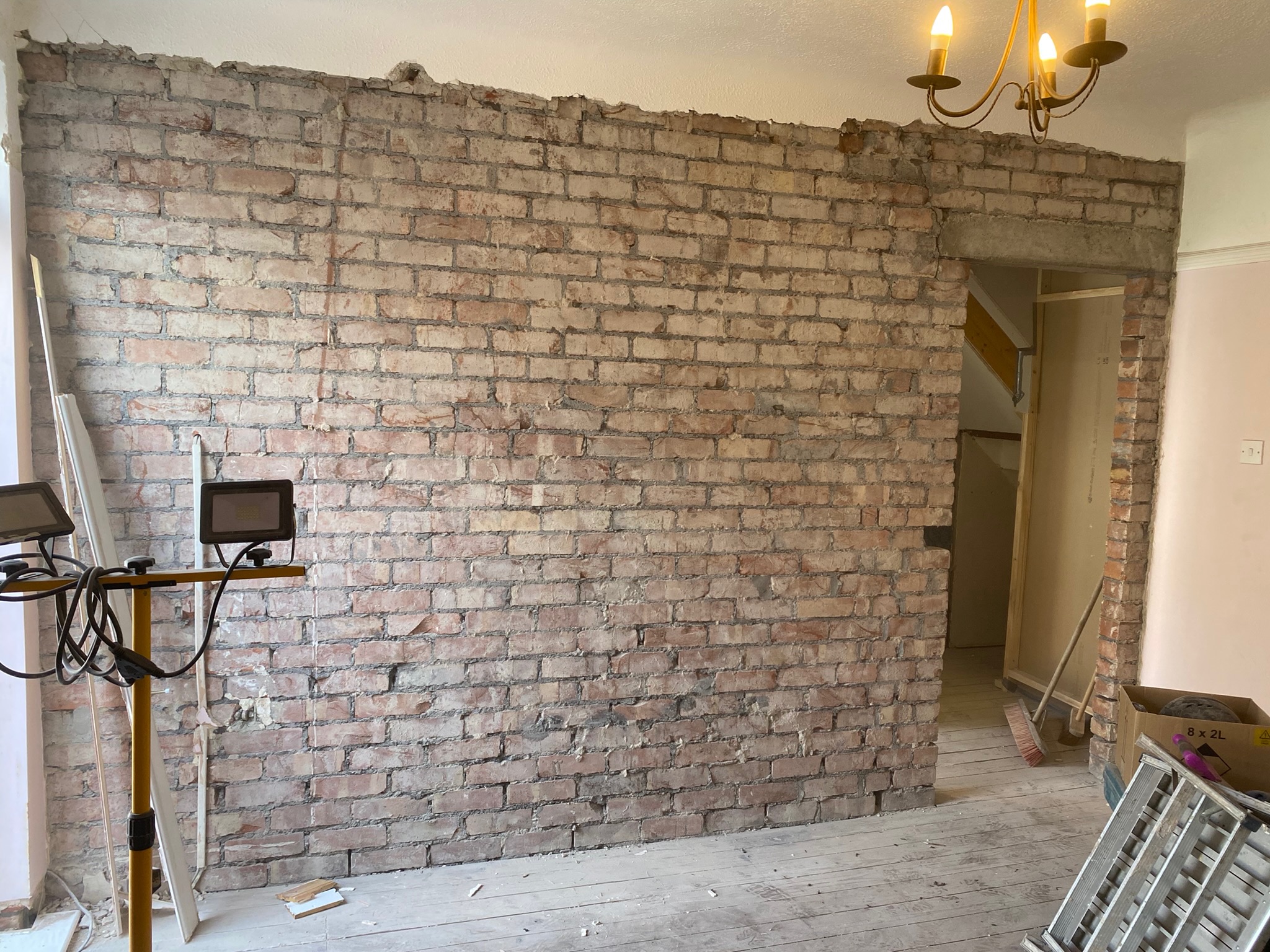 Hummingbird Bathrooms and Kitchens renovation project featuring a bare brick wall, open doorway to the right, construction lights to the left, exposed floorboards, various building tools, and a chandelier overhead—expert bathroom, kitchen and home design services in the UK.