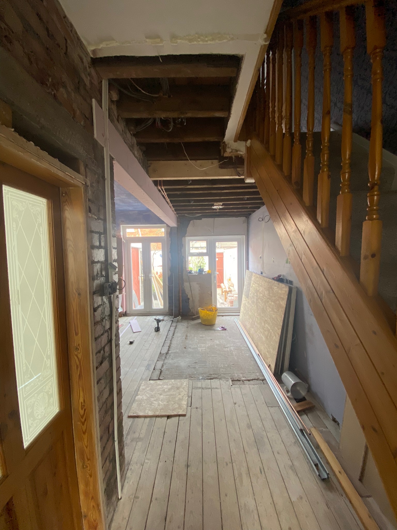Home renovation in progress by Hummingbird Bathrooms and Kitchens, showcasing exposed wooden floors, unfinished walls, building materials, and staircase on the right. Natural sunlight brightens the space through glass doors at the back. Specialist in UK kitchen, bathroom design and construction services.