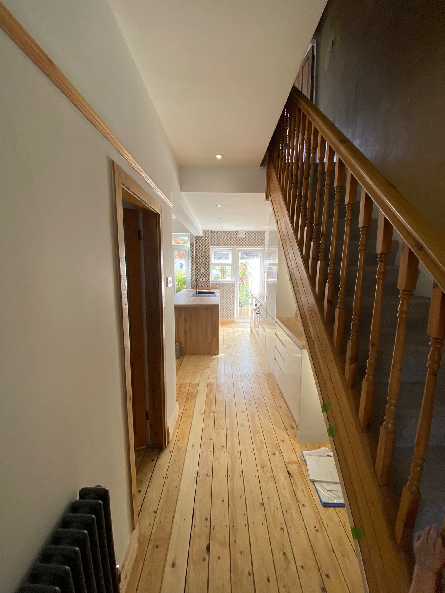 Modern hallway with wooden floors leading to a contemporary kitchen, featuring a staircase with wooden bannisters on the right. Natural light fills the space through glass doors at the end. Interior design by Hummingbird Bathrooms and Kitchens, specialists in premium UK kitchen and bathroom refurbishments, construction services, and bespoke design solutions.