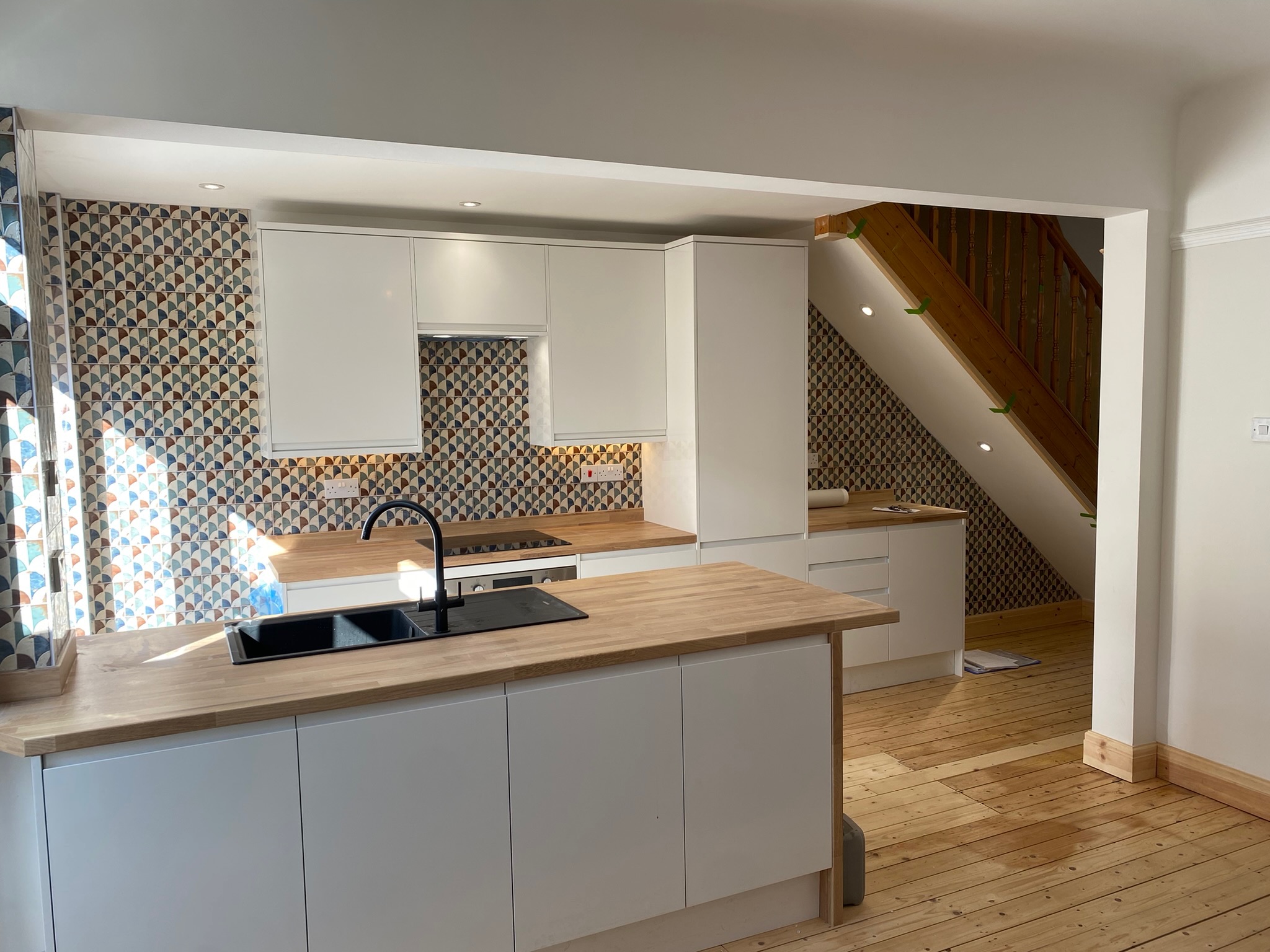 Contemporary kitchen design by Hummingbird Bathrooms and Kitchens, featuring sleek white cabinetry, wooden worktops, stylish patterned tiled splashback, and a modern black sink. Wooden staircase with bannisters visible in the background and natural sunlight highlighting this bespoke UK kitchen renovation.