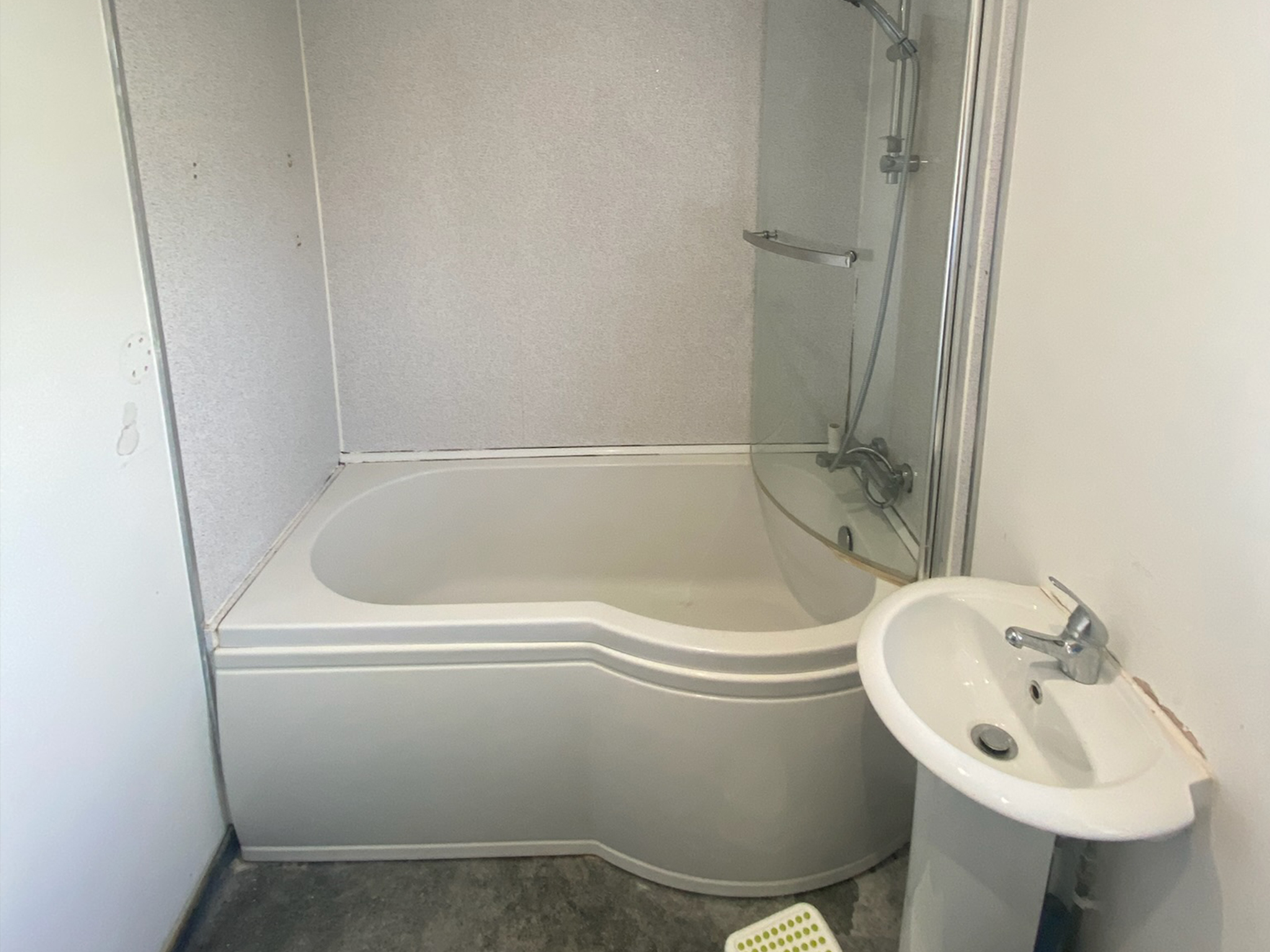 Small bathroom before it was upgraded by Hummingbird Bathrooms and Kitchens, showcasing a white bath with glass shower screen, wall-mounted shower, compact white basin with silver tap, plain white walls, and stylish grey floor—expert bathroom design and installation in the UK.