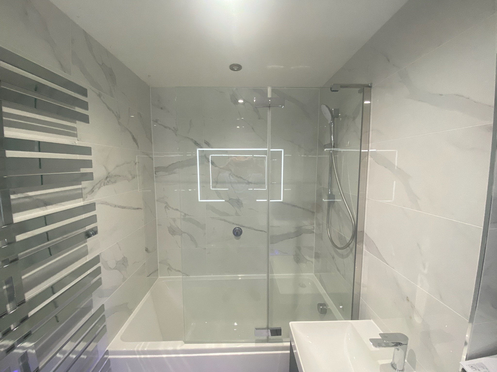 Modern bathroom designed by Hummingbird Bathrooms and Kitchens, featuring white marble-style tiles, a sleek bath with glass shower screen, overhead shower, chrome heated towel rail, and compact white basin. The space is brightly lit with natural light reflecting off the glossy tiles—expertly fitted by our UK construction and bathroom design specialists.
